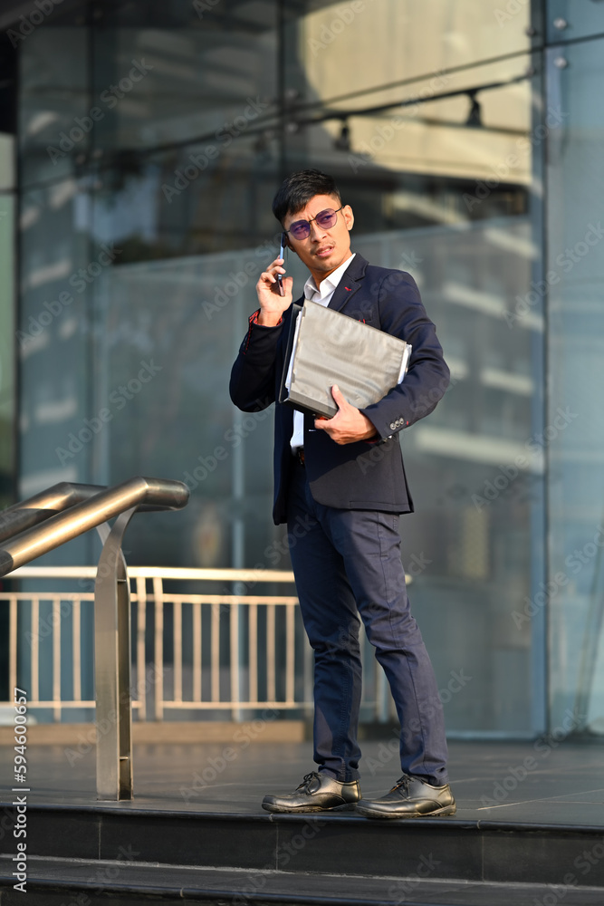 Full length portrait of adult businessman in formalwear standing outside building and talking on mobile phone Full length portrait of adult businessman in formalwear standing outside building and talking on mobile phone