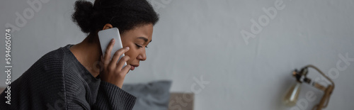 Upset multiracial woman in sweater talking on cellphone in bedroom at home, banner.