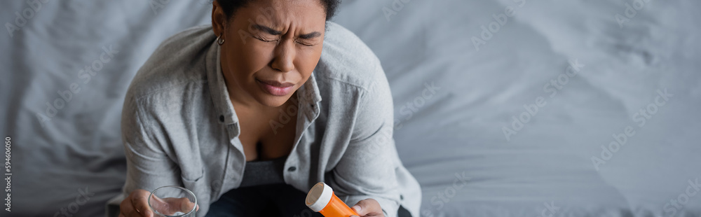 Φωτογραφία High angle view of depressed multiracial woman holding water and antidepressant pills on bed at home, banner