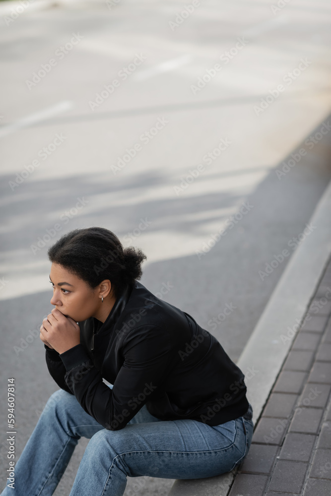 Young multiracial woman with depression looking away while sitting on border outdoors.