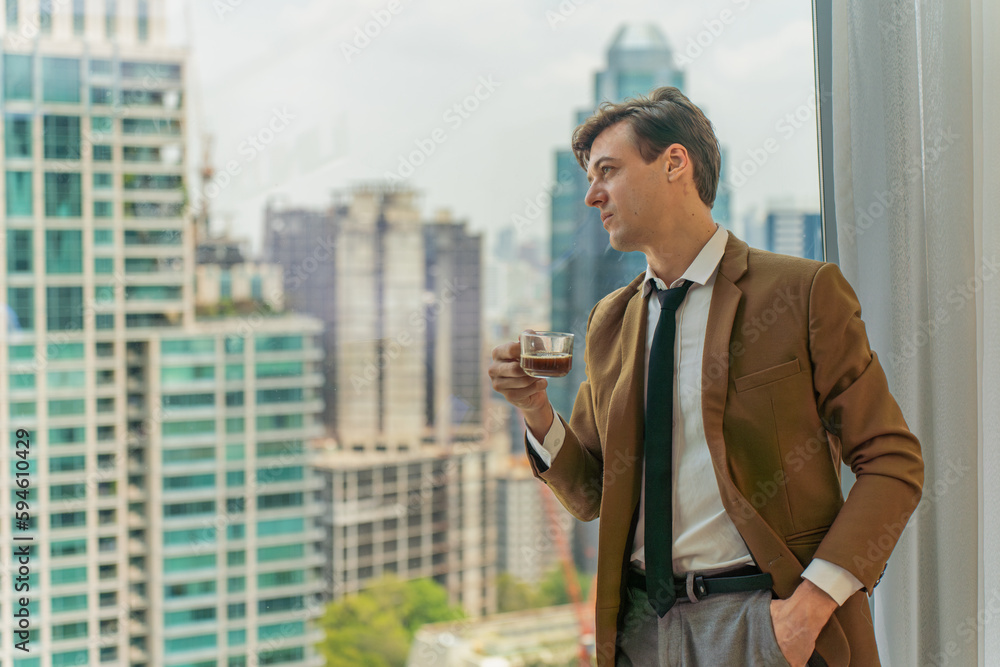 Handsome young white businessman holding a cup of coffee looking out the window view of city skyline in a business district area