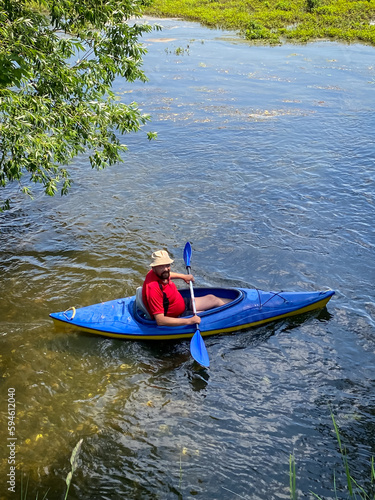 A large man is sitting on the river in a kayak, he has an oar in his hand, he is in a hat and a red T-shirt.