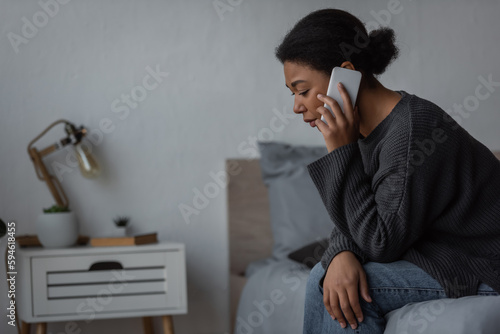 heartbroken multiracial woman in sweater talking on smartphone on bed at home.