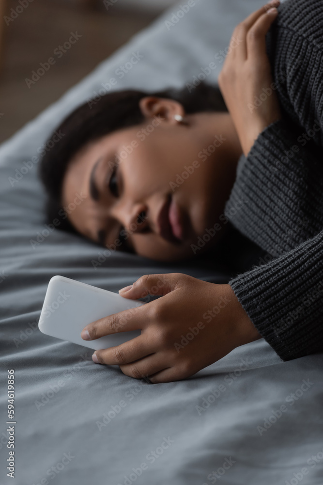 Blurred multiracial woman with depression using smartphone while lying on bed at home. Blurred multiracial woman with depression using smartphone while lying on bed at home.