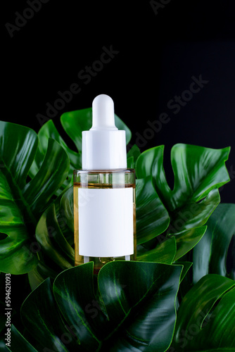 Transparent dropper bottle on black background with monstera palm leaf. Blank aromatic oil container design, medical packaging template. Herbal cosmetic with vitamin c. Flat lay, top view..