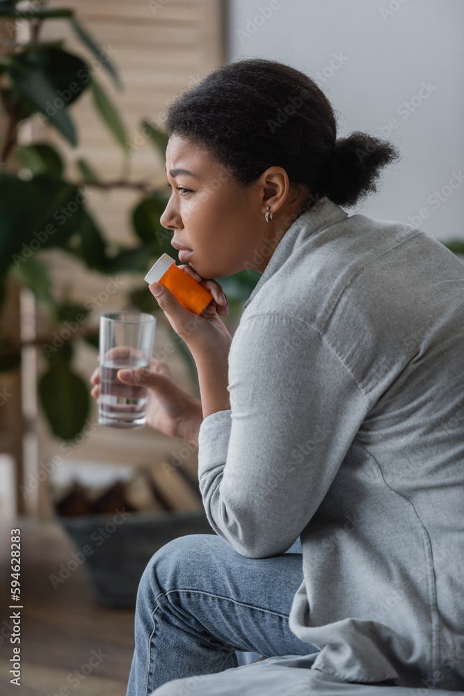 Fototapeta premium Side view of sad multiracial woman holding pills and water while sitting on bed at home.
