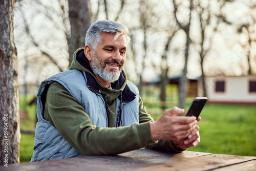 A smiling senior man uses his mobile phone and sits on a park bench.