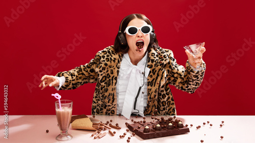Foto Emotional, eccentric woman in sunglasses and animal print coat, dj on party, playing music and eating chocolate against red background