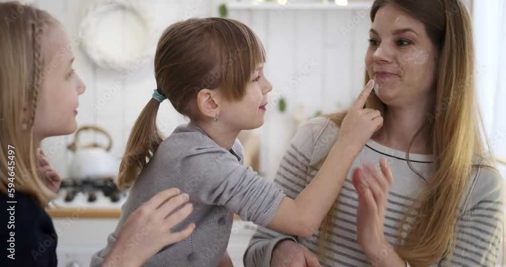 Slow motion shot of playful daughters applying flour on young mother's face while making cookies in kitchen during weekend Slow motion shot of playful daughters applying flour on young mother's face while making cookies in kitchen during weekend