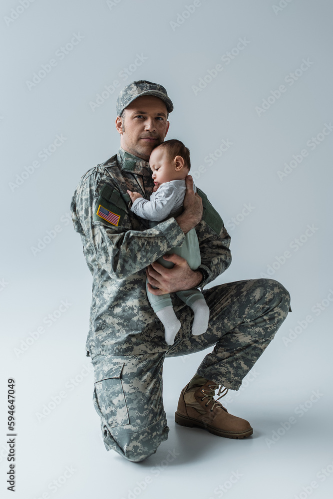 US soldier in uniform holding in arms newborn boy and looking at camera on grey. US soldier in uniform holding in arms newborn boy and looking at camera on grey.