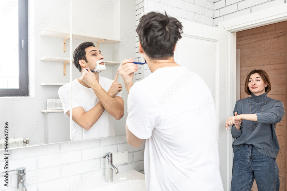 Young handsome man shaving in front of the mirror in the bathroom and his wife waiting for him on the door Young handsome man shaving in front of the mirror in the bathroom and his wife waiting for him on the door