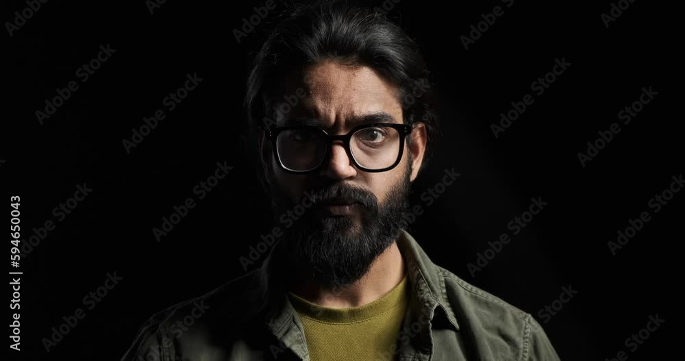 Slow motion shot of angry young man with eyeglasses and beard staring seriously at camera isolated against black background Slow motion shot of angry young man with eyeglasses and beard staring seriously at camera isolated against black background