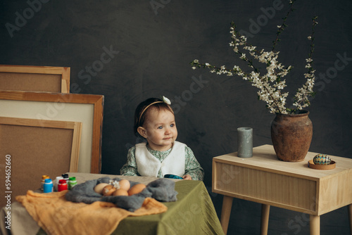 Little cute girl with paints and eggs in a studio with beautiful decorations and spring flowers and picture frames