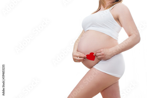 Portret nierozpoznawalnej kobiety podczas ostatnich miesięcy ciąży, trzymającej swój duży brzuch i czerwone serduszko delikatnie stojącej przy ścianie na białym tle photo