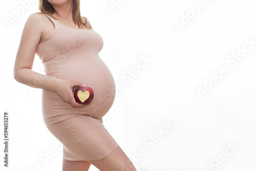 Portret strzał młodej pięknej kobiety w trzecim trymestrze ciąży. Zbliżenie ciężarnej kobiety z rękami na okrągłym brzuchu. Oczekiwanie koncepcji dziecka. Tło, miejsce na kopię, jabłko z wyciętym serc