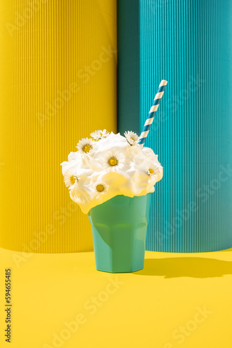 Creamy milkshake with daisy flowers and paper straw on yellow and blue background.