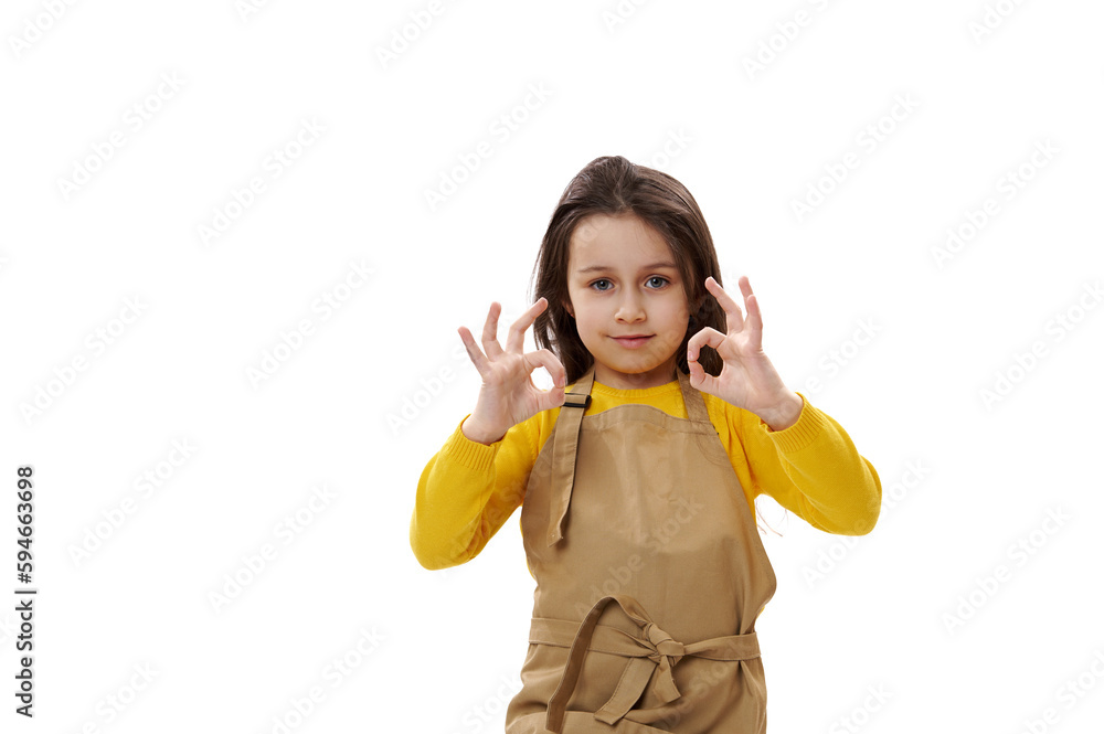 Caucasian little child girl in beige chef's apron, gesturing with OK sign, expressing agreement and apreciation on white isolated background. Copy ad space. Cooking class. Food and drink consumerism Caucasian little child girl in beige chef's apron, gesturing with OK sign, expressing agreement and apreciation on white isolated background. Copy ad space. Cooking class. Food and drink consumerism
