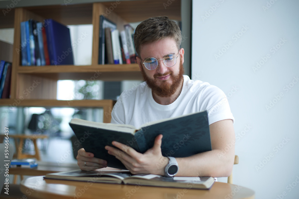 Portrait of young bearded university or college happy student in glasses sitting in library and read book. Education concept. Portrait of young bearded university or college happy student in glasses sitting in library and read book. Education concept.