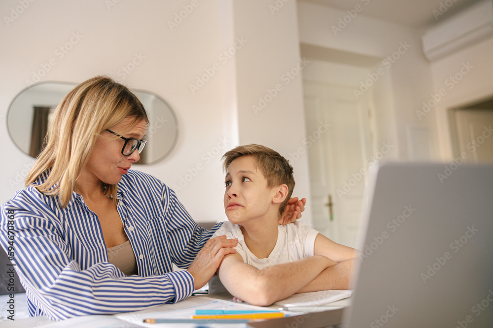 Tired sad kid sits at table with laptop and textbooks while mom help him with homework Tired sad kid sits at table with laptop and textbooks while mom help him with homework
