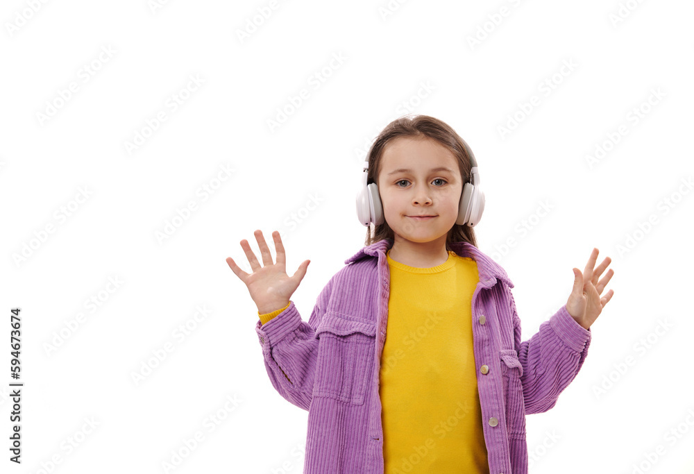 Caucasian happy adorable little girl in bright yellow sweater and purple jacket, wearing wireless headphones, looks and camera and shows her hands palms up, isolated over white background. Copy space Caucasian happy adorable little girl in bright yellow sweater and purple jacket, wearing wireless headphones, looks and camera and shows her hands palms up, isolated over white background. Copy space