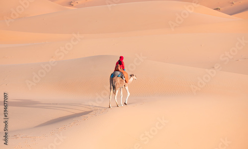 A woman in a red turban riding a camel across the thin sand dunes of the in Western Sahara Desert  Morocco  Africa