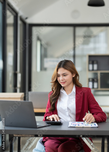 Beautiful asian businesswoman working on laptop and calculating with financial calculator from statistics data graphs, charts. Successful business results in modern office with joy.