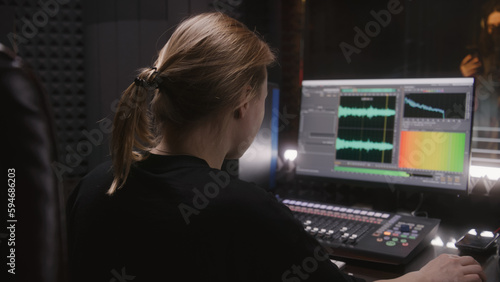 Male singer records song in soundproof room. Female audio engineer, producer uses control surface. Computer screen showing DAW software user interface with song tracks. Work in sound recording studio.
