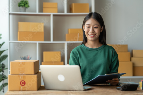 starting a small business SME, Asian businesswoman or e-commerce store owner using laptop and chart to check orders, online ordering, shipping tracking number to prepare packages in office.