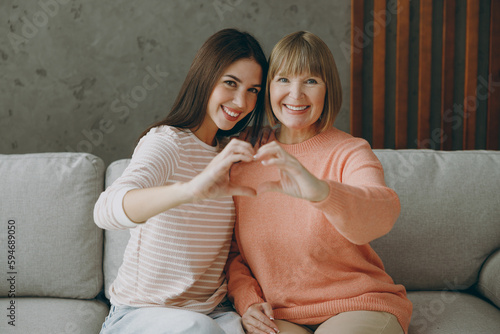 Two adult women mature mom young kid wear casual clothes show shape heart with hand heart-shape sign hug sit on gray sofa stay home flat rest relax spend free spare time in living room Family concept