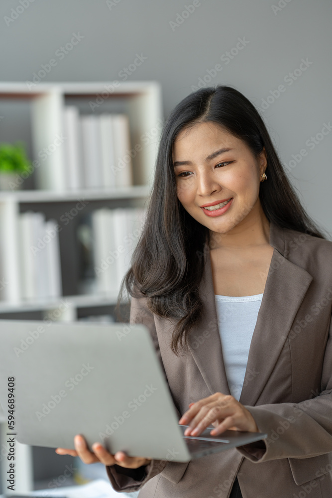 Asian businesswoman smiling confidently successful entrepreneur in the financial business professional company executive Wearing suit standing holding laptop searching business information in office Asian businesswoman smiling confidently successful entrepreneur in the financial business professional company executive Wearing suit standing holding laptop searching business information in office