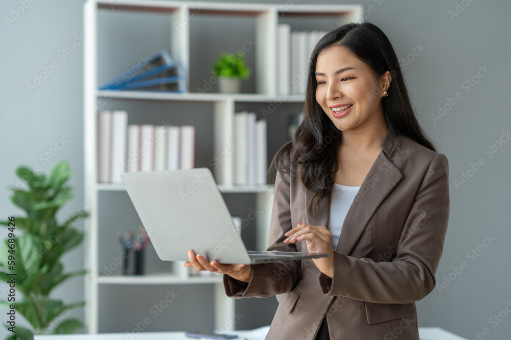 Asian businesswoman smiling confidently successful entrepreneur in the financial business professional company executive Wearing suit standing holding laptop searching business information in office Asian businesswoman smiling confidently successful entrepreneur in the financial business professional company executive Wearing suit standing holding laptop searching business information in office