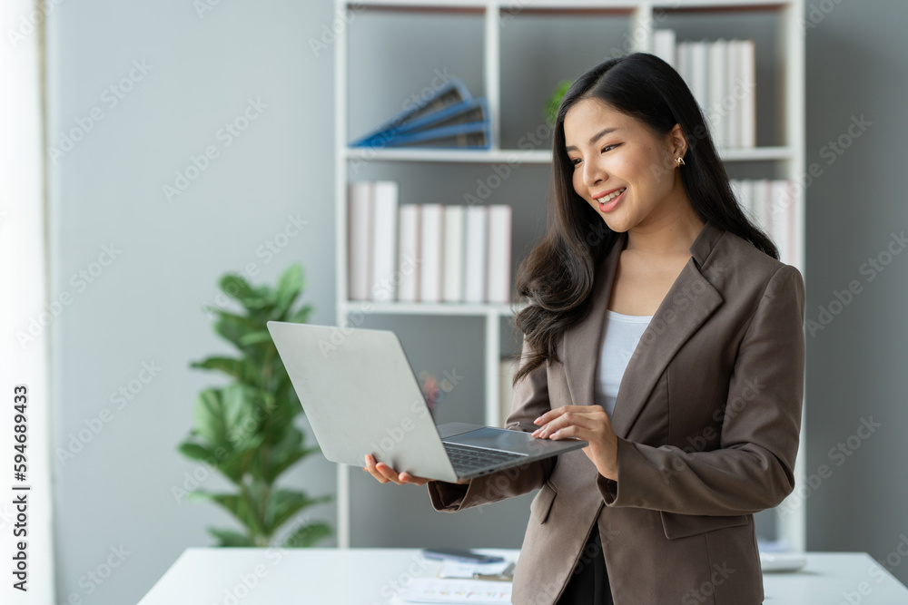 Asian businesswoman smiling confidently successful entrepreneur in the financial business professional company executive Wearing suit standing holding laptop searching business information in office Asian businesswoman smiling confidently successful entrepreneur in the financial business professional company executive Wearing suit standing holding laptop searching business information in office