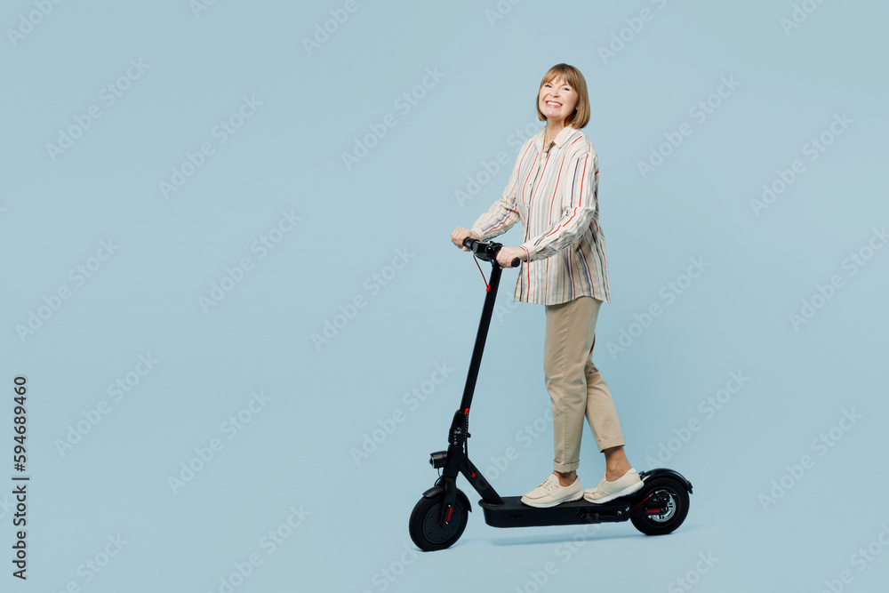 Full body side view smiling happy fun elderly woman 50s years old wear shirt riding e-scooter look camera isolated on plain pastel light blue cyan color background studio portrait. Lifestyle concept. Full body side view smiling happy fun elderly woman 50s years old wear shirt riding e-scooter look camera isolated on plain pastel light blue cyan color background studio portrait. Lifestyle concept.