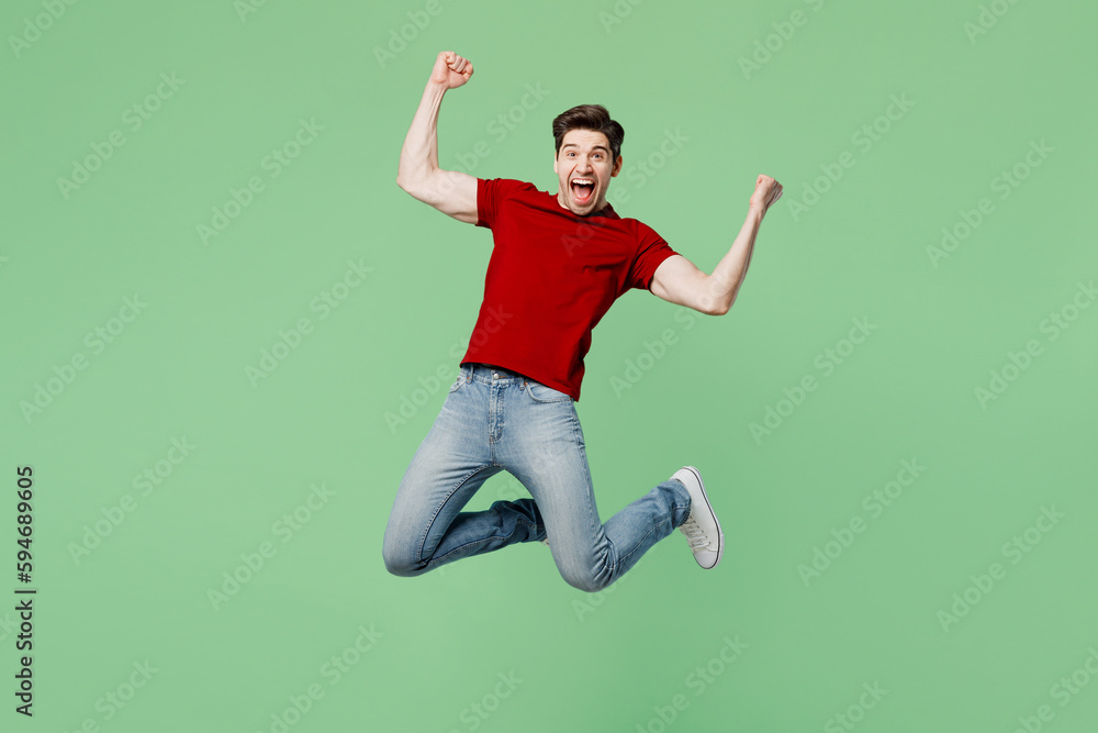 Full body young fun brunet caucasian man he wears red t-shirt casual clothes jump high do winner gesture clench fist isolated on plain pastel light green background studio portrait. Lifestyle concept.