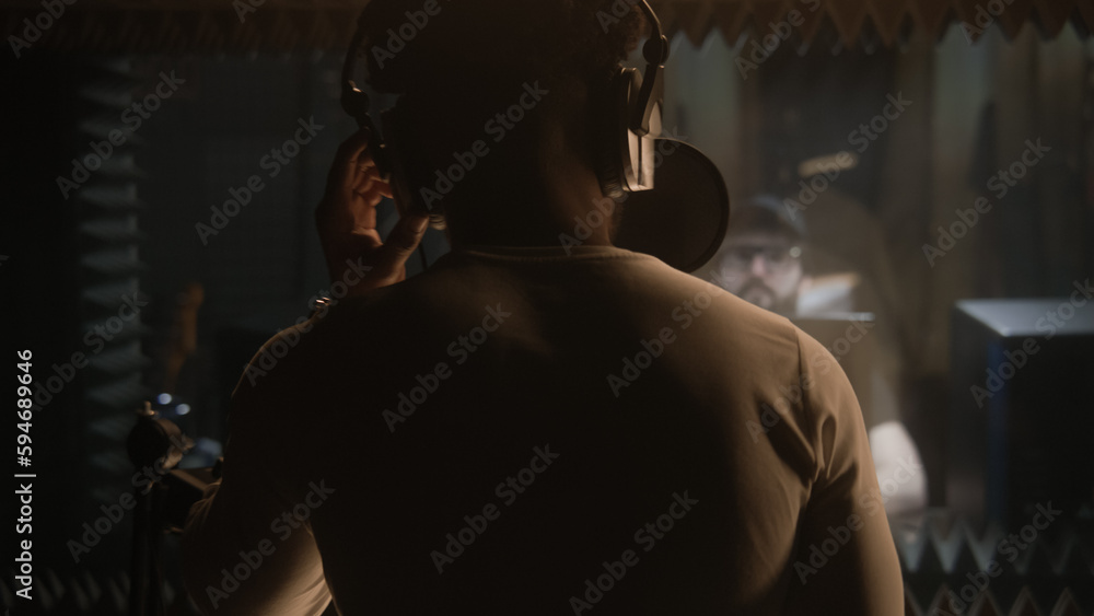 Naklejka premium African American vocalist sings composition into microphone in soundproof room. Experienced audio engineer or producer works with singer in sound recording studio. Music production concept. Back view.