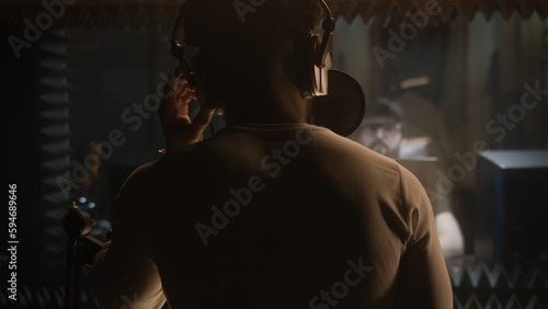 African American vocalist sings composition into microphone in soundproof room. Experienced audio engineer or producer works with singer in sound recording studio. Music production concept. Back view. © Framestock