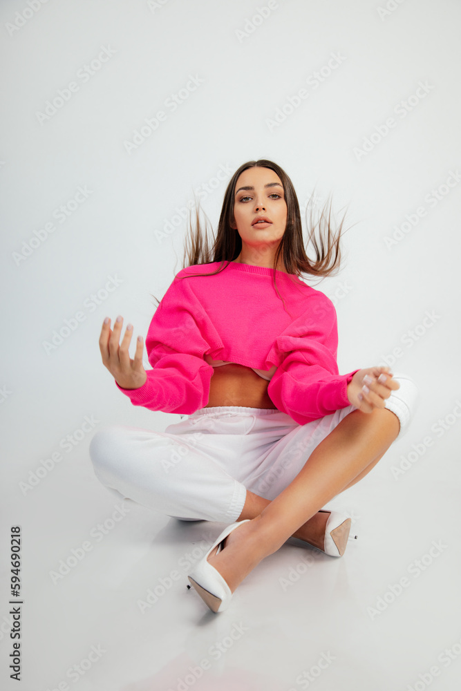 Stylish woman in pink crop pullover, pants and high heel shoes sitting on floor with flying hair. Sport casual wear. Stylish woman in pink crop pullover, pants and high heel shoes sitting on floor with flying hair. Sport casual wear.