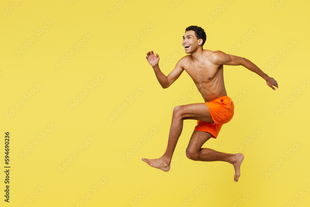 Full body young fun sporty man wears orange shorts swimsuit relax near hotel pool jump high look aside on area run fast isolated on plain yellow background. Summer vacation sea rest sun tan concept.