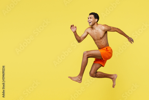 Full body young fun sporty man wears orange shorts swimsuit relax near hotel pool jump high look aside on area run fast isolated on plain yellow background. Summer vacation sea rest sun tan concept. © ViDi Studio
