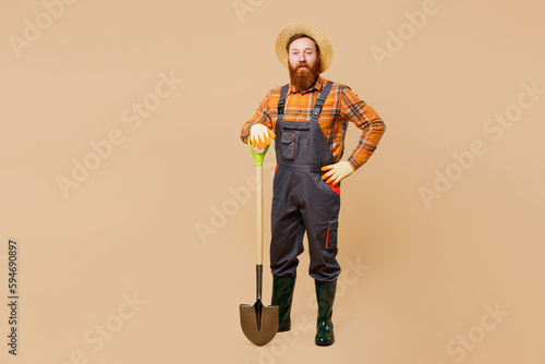 Full body happy young bearded man wears straw hat overalls work in garden hold shovel digging look camera isolated on plain pastel light beige color background studio portrait. Plant caring concept.