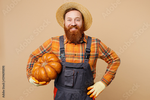 Happy smiling cheerful young bearded man wear straw hat overalls work in garden hold pumpkin stand akimbo isolated on plain pastel light beige color background studio portrait. Plant caring concept.