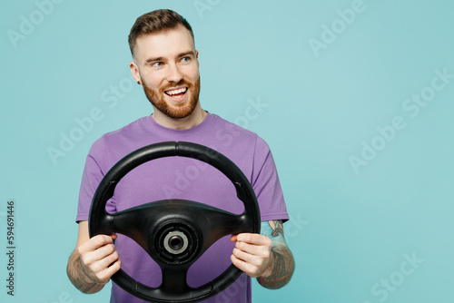 Young smiling cheerful fun man he wear purple t-shirt hold steering wheel driving car look aside on area isolated on plain pastel light blue cyan color background studio portrait. Lifestyle concept.
