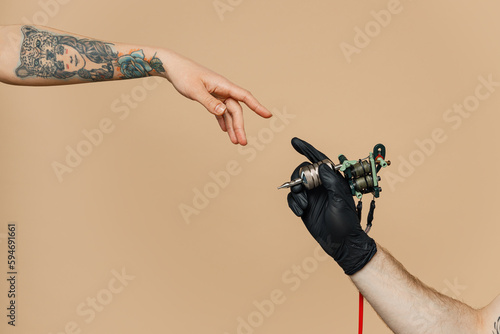 Close up cropped photo of tattooer master artist male hold in hand machine black ink in jar, equipment for making tattoo art on body, touch female finger arm isolated on pastel plain beige background. photo