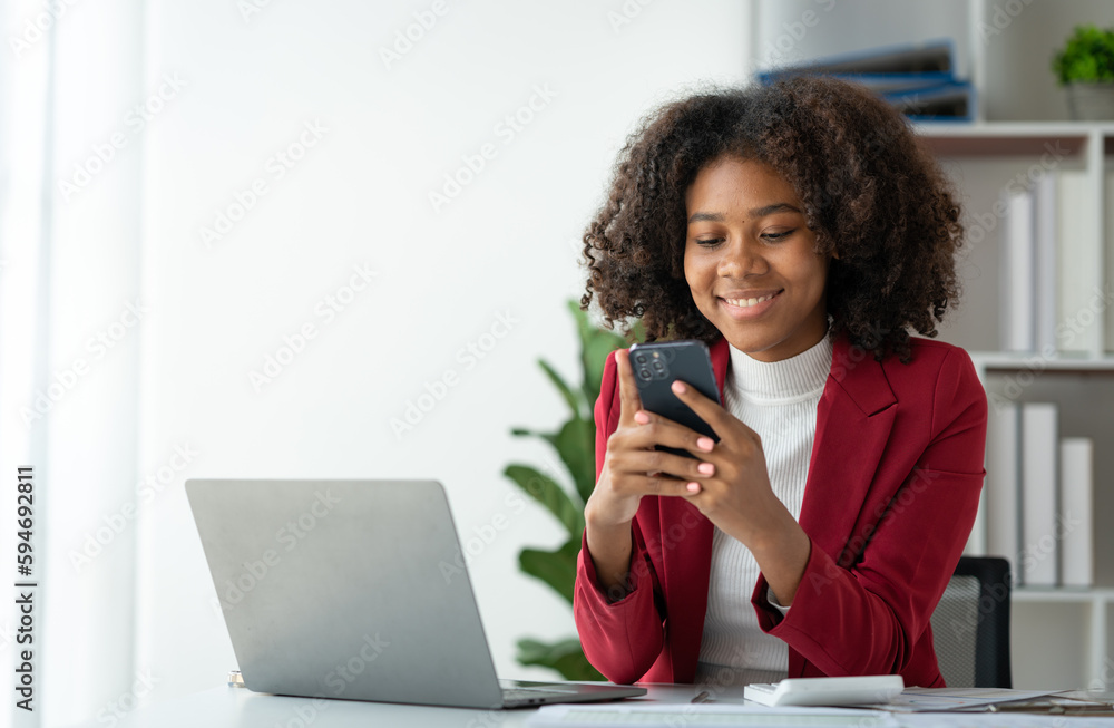 African American business woman holding a mobile phone to read financial business news Online marketing on mobile apps to process emails in meetings sitting at laptop desk in office. African American business woman holding a mobile phone to read financial business news Online marketing on mobile apps to process emails in meetings sitting at laptop desk in office.