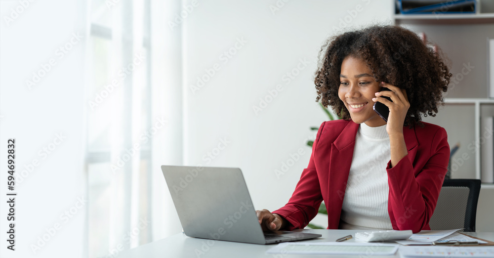 African business woman American chatting on cell phone happily chatting and reporting earnings to clients or investors working on a laptop in the office. African business woman American chatting on cell phone happily chatting and reporting earnings to clients or investors working on a laptop in the office.