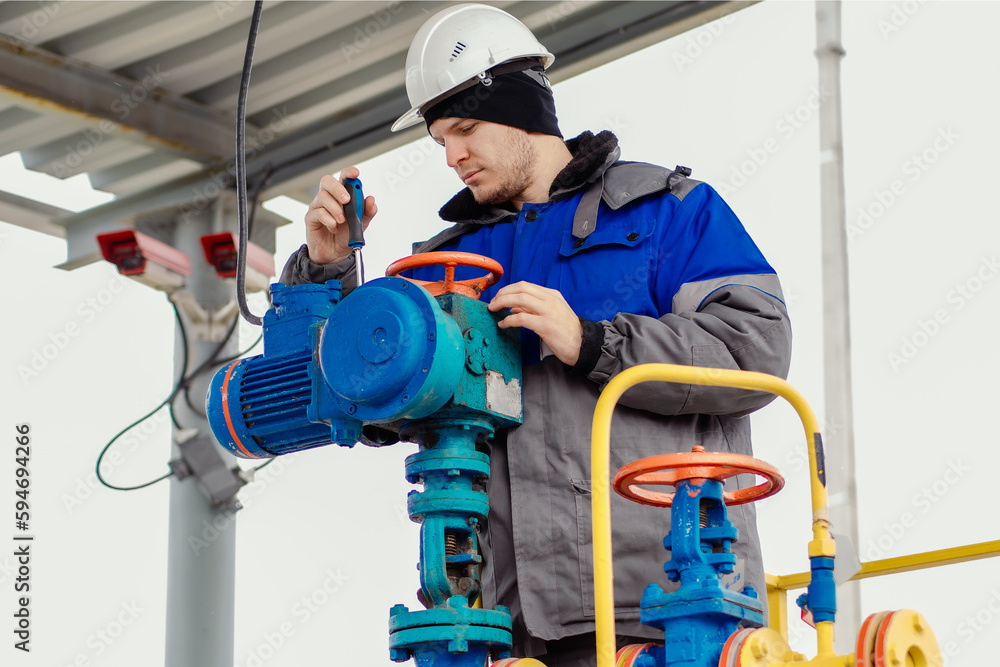 Fototapeta premium An engineer with a screwdriver repairs equipment at a gas station. Male engineer in helmet and winter clothing doing maintenance on industrial gas equipment outside..