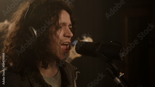 Close up shot of male singer singing lyrical composition into microphone in soundproof room. Vocalist in professional headphones records song in sound recording studio. Concept of music production.