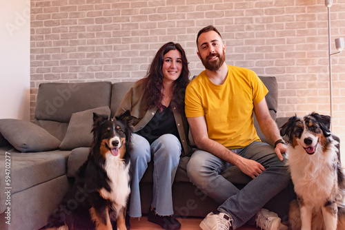 Young couple sitting on the couch at home with their two dogs sitting on the floor. They are all looking at the camera. Australian Sheepdog,