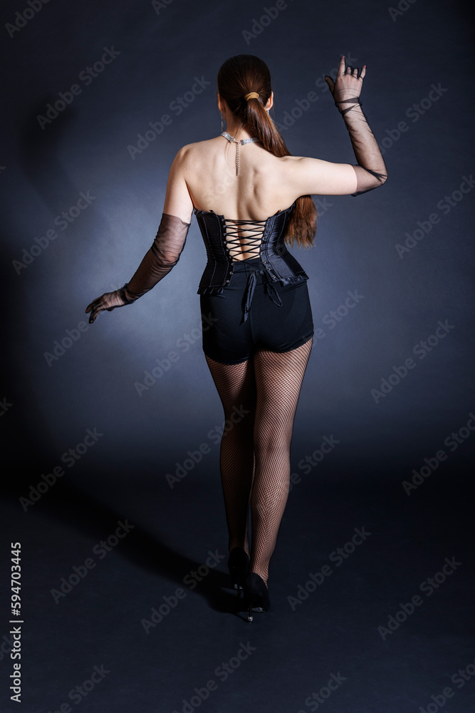 A seductive young woman in a black corset poses elegantly against a dark background, standing with her back to the viewer.