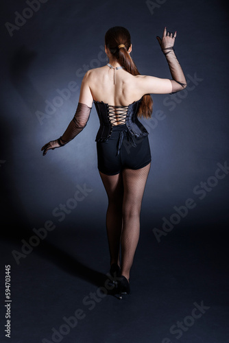 A seductive young woman in a black corset poses elegantly against a dark background, standing with her back to the viewer.